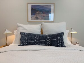 A bed with white bedding and two decorative pillows, one with a leaf pattern, in a room with a framed picture on the wall. - Modern Denver Suite (Denver)