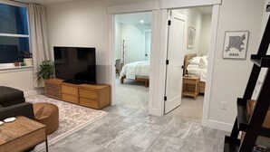 A modern living room with a flat-screen TV, a sofa, a coffee table, and a rug. There is a bedroom visible through an open door with a bed and bedside table.