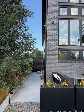 A modern building with a brick facade, large windows, and a garden area with planters and outdoor furniture.