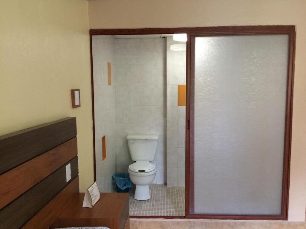 Casa de banho com sanita, um banco e porta que dá para outro espaço.