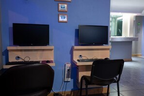 A room with two monitors, a desk, a chair, and a small shelf with framed pictures. - Hotel Fabiola (Mexico City)