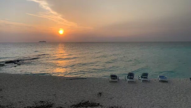 Ein Strand mit Sonnenuntergang, ruhiger See und vier Liegestühlen.