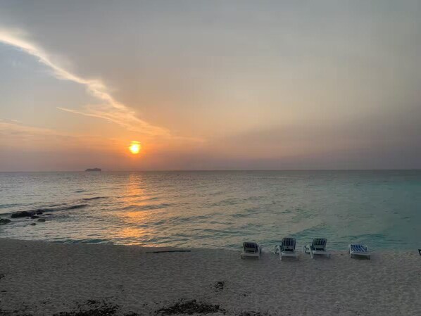 A beach with a sunset, calm sea, and four lounge chairs.