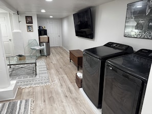 A laundry room with two black washers and dryers, a glass table with books, a wicker chair, and a white door.