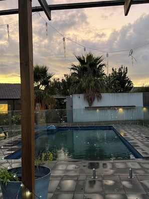 Piscina com deck ladrilhado, cercada por palmeiras e um prédio com telhado inclinado.