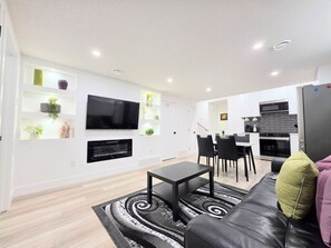 A modern living room with a flat-screen TV, a fireplace, a dining area with chairs, and a kitchen with a microwave. - New 2-Bed Suite in SW Edmonton near Windermere & YEG Airport (Edmonton)