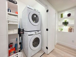 A compact laundry room with two stacked washing machines, a drying rack, and a shelf with cleaning supplies. - New 2-Bed Suite in SW Edmonton near Windermere & YEG Airport (Edmonton)