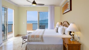 Quarto de hotel com cama, luminárias de cabeceira, mesa de cabeceira, ventilador de teto, varanda com vista para o mar e um quadro na parede.