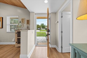 A hallway with a wooden door, a window with blinds, and a small cabinet with decorative items. - 4BR Heated Pool Near Beach | Good Morning Rentals (Jacksonville Beach)