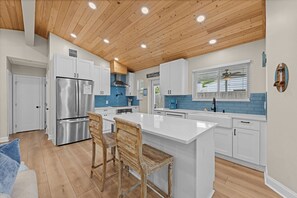 A modern kitchen with white cabinets, a stainless steel refrigerator, wooden ceiling, and a large island with bar stools. - 4BR Heated Pool Near Beach | Good Morning Rentals (Jacksonville Beach)