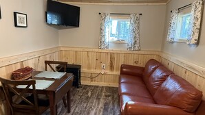 A room with a wooden ceiling, a brown leather sofa, a dining table with chairs, a flat-screen TV mounted on the wall, and two windows with floral curtains.