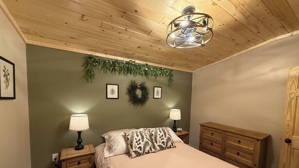 A bedroom with a bed, two nightstands with lamps, a wooden dresser, a ceiling fan, and a wall with a wreath and framed leaves.