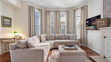 A corner living room with a beige sectional sofa, a small table, and a TV mounted on the wall.