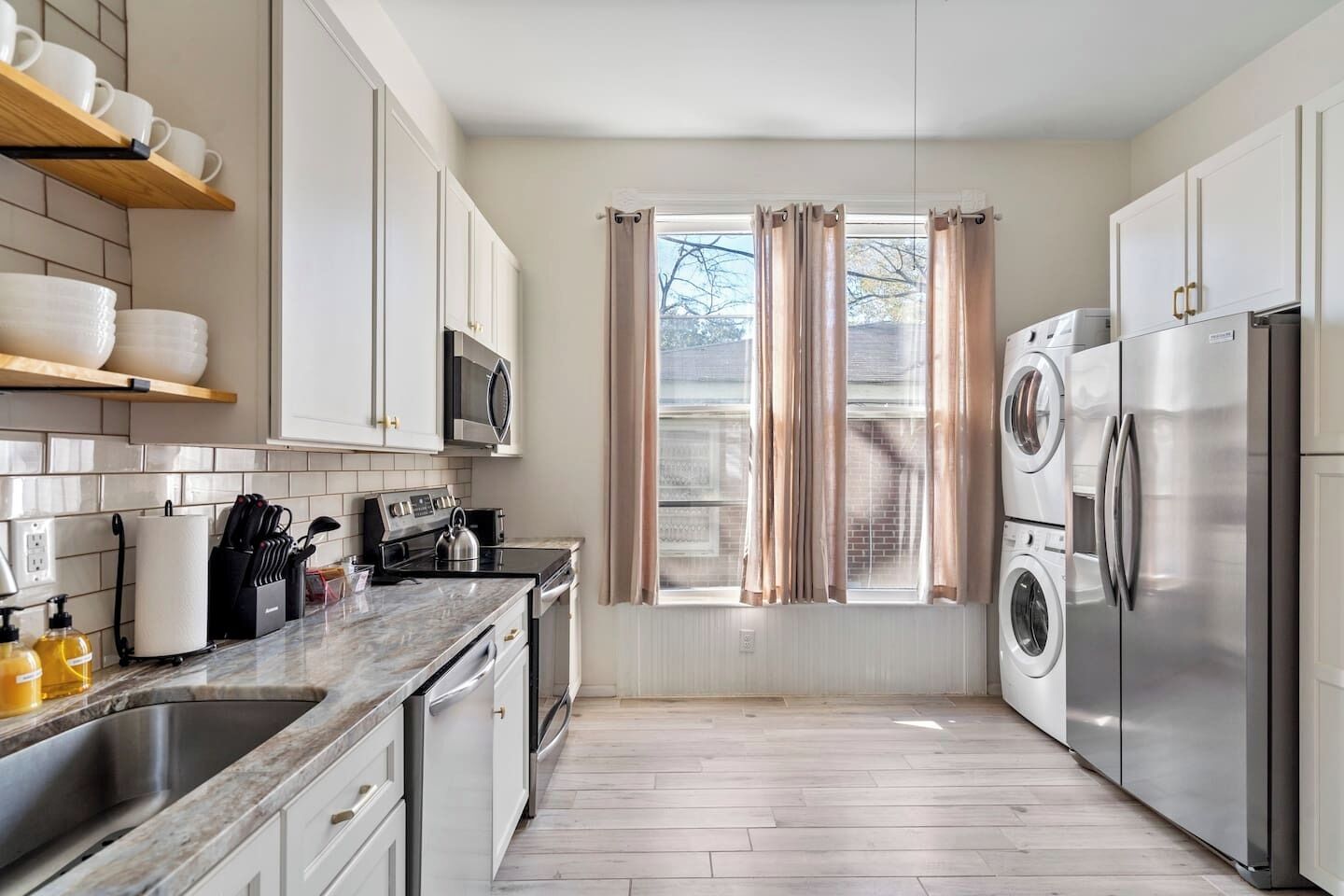 A modern kitchen with a stainless steel refrigerator, microwave, and oven, wooden cabinets, and a window with curtains.