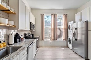 A modern kitchen with a stainless steel refrigerator, microwave, and oven, wooden cabinets, and a window with curtains. - 1889 Historic Gem · Walk to Riverwalk & Downtown (Wilmington)