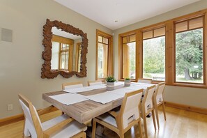 A dining room with a wooden table, white chairs, a large mirror, and large windows with a view of trees. - Cozy ski chalet on Nicklaus North Golf Course (Whistler)