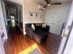 A hallway with wooden flooring, a gray sofa with cushions, a ceiling fan, and a white door. - Cozy 2-bedroom house in pleasant Apalachicola with AC (Apalachicola)