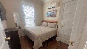 Ein Schlafzimmer mit Bett, Kommode, Fenster mit Jalousien, Klimaanlage und Tür.