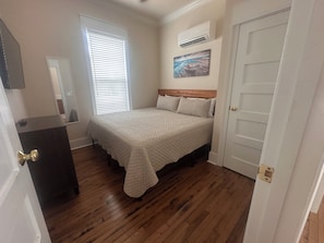 Ein Schlafzimmer mit Bett, Kommode, Fenster mit Jalousien, Klimaanlage und Tür.