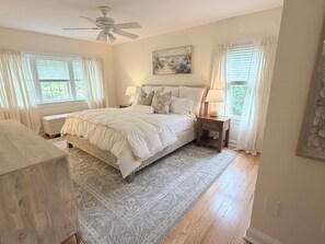 Um quarto com cama, mesas de cabeceira, ventoinha no teto e janela com cortinas.