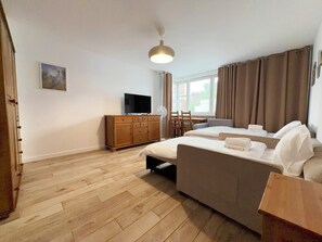 Ein Hotelzimmer mit Holzboden, einem Fernseher auf einer Holzkommode, einem Bett mit weißer Bettwäsche, einem Schreibtisch mit Stuhl und einem Fenster mit Vorhängen.