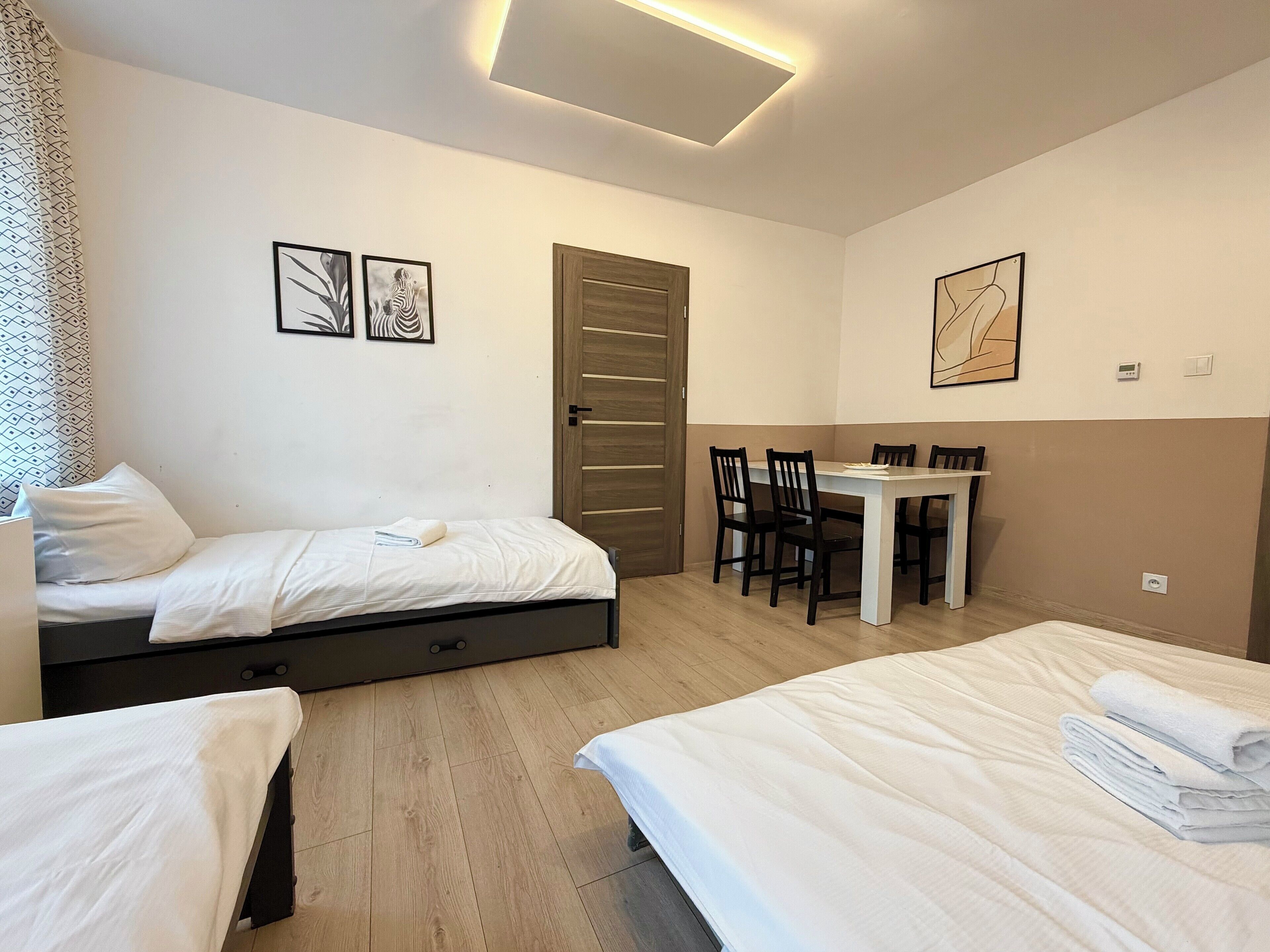 A hotel room with two beds, a dining table with chairs, a wooden door, and artwork on the walls.