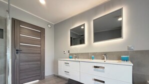 A modern bathroom with a glass shower enclosure, a double vanity with a large mirror, and a dark wooden door. - Park Leśnicki Homely Studio #2 (Wrocław)