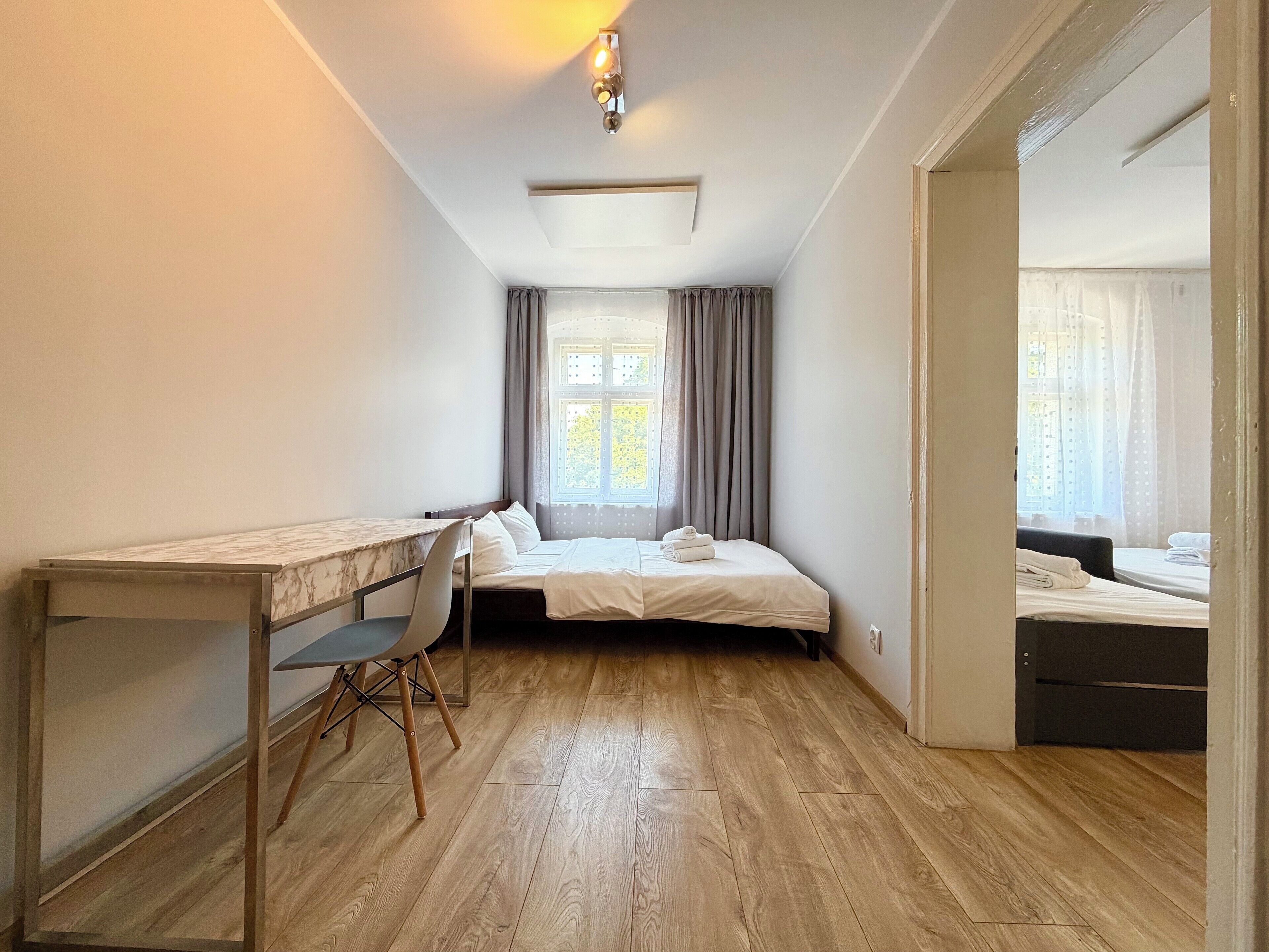 Une chambre à coucher avec un lit, un bureau et une chaise. Il y a une fenêtre avec des rideaux et une porte qui donne sur une autre pièce.