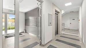 Corredor moderno de hotel com porta de vidro, elevador embutido na parede e piso ladrilhado.