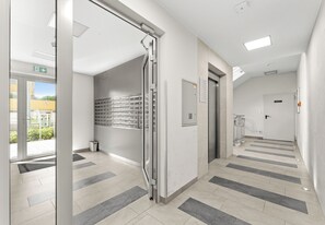 Un pasillo moderno de hotel con puerta de vidrio, un ascensor empotrado en la pared y piso de baldosas.