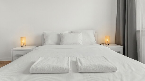 Cama bem arrumada com lençóis brancos, duas luminárias de cabeceira e uma janela com cortinas.