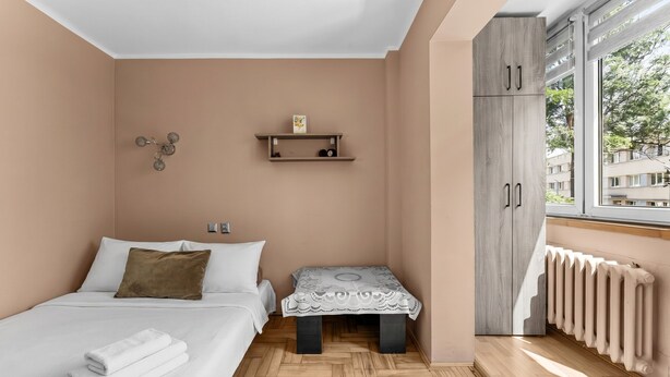 Quarto com cama, mesa de cabeceira, prateleira na parede, radiador e janela com vista.