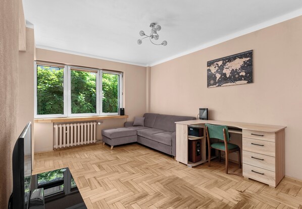 A room with a large window, a grey sofa, a wooden desk with drawers, a chair, and a radiator. - Sołtyka Comfort Apartment (Kraków)