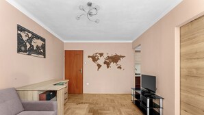 Um quarto com piso de madeira em padrão herringbone, um sofá cinza, uma escrivaninha de madeira, um adesivo de parede com um mapa mundi, um ventilador de teto e um rack para TV.