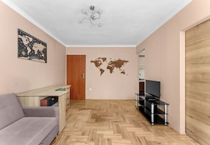 Ein Zimmer mit Fischgrätparkett, einem grauen Sofa, einem Holztisch, einer Wandsticker-Weltkarte, einem Deckenventilator und einem TV-Möbel.