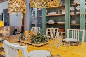 Ein Essbereich mit einem Holztisch, gedeckt für eine Mahlzeit, umgeben von weißen Stühlen, mit gewobenen Leuchten und einer Vitrine mit Geschirr.