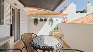 En balkon med et rundt bord og fletstole, en hvid væg med plantekrukker, og et tagtækket tag.
