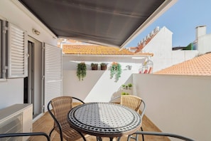 Ein Balkon mit rundem Tisch und Korbstühlen, eine weiße Wand mit Topfpflanzen und ein Ziegeldach.