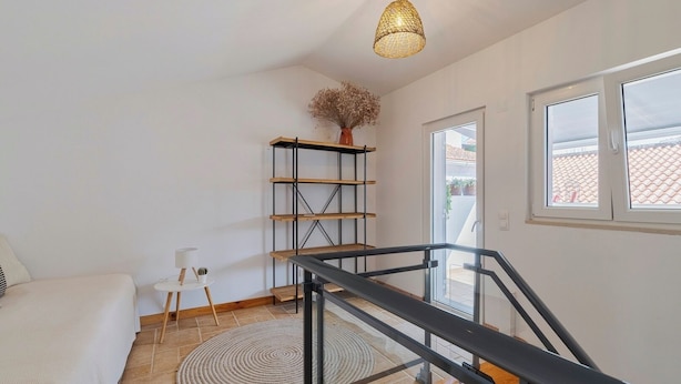Et soveværelse med en seng, et lille bord med lampe, en fletvæglampe, en træhylde og en trappe.