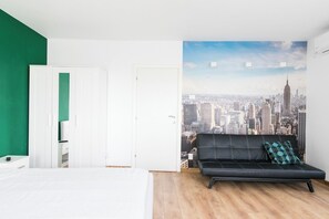Ett rum med en säng, en grön vägg, en vit dörr, en svart soffa med en mönstrad kudde och en väggmålning som föreställer en stadsbild.