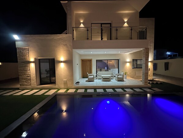 Ein modernes, zweistöckiges Haus mit Swimmingpool, Sitzgelegenheiten im Freien und einer gut beleuchteten Eingangsparte.