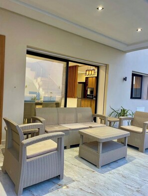 A modern living room with wicker furniture, a glass sliding door, and a kitchen visible through the door. - Discover our magnificent Cercina villa, nestled in the heart of the tourist area (Sfax)