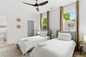Un dormitorio con dos camas, un ventilador de techo, una ventana con cortinas y vista a una casa.
