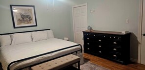 A bedroom with a bed, a dresser, a bench, a framed picture, and a door. - Pondview Gardens (Grand Falls-Windsor)