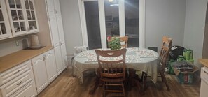 A dining area with a table set for two, surrounded by wooden chairs, a sewing machine, and a window with a view of the kitchen. - Pondview Gardens (Grand Falls-Windsor)