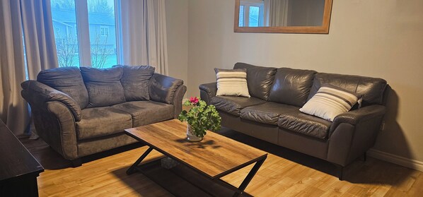 Two leather sofas with cushions, a wooden coffee table with a plant, and a large window with curtains. - Pondview Gardens (Grand Falls-Windsor)
