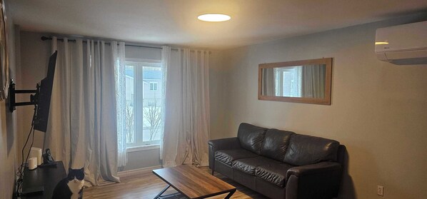 Un salon avec un canapé en cuir noir, une table basse en bois, un téléviseur à écran plat fixé au mur, une fenêtre avec des rideaux et un appareil de climatisation.