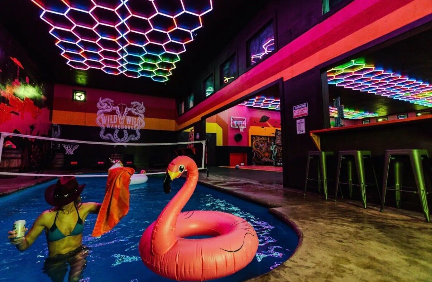 Een zwembadgedeelte met een grote opblaasflamingo, een volleybalnet en een neonverlicht plafond.