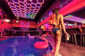 A pool area with a neon-lit ceiling, a flamingo float, and a basketball hoop.