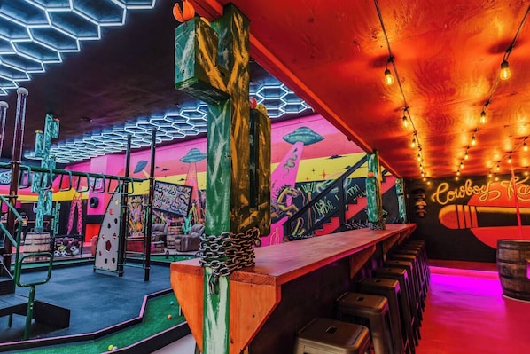 Un espace bar animé, illuminé de néons, avec un comptoir en bois et une fresque murale représentant des œuvres d’art sur le thème de l’espace.